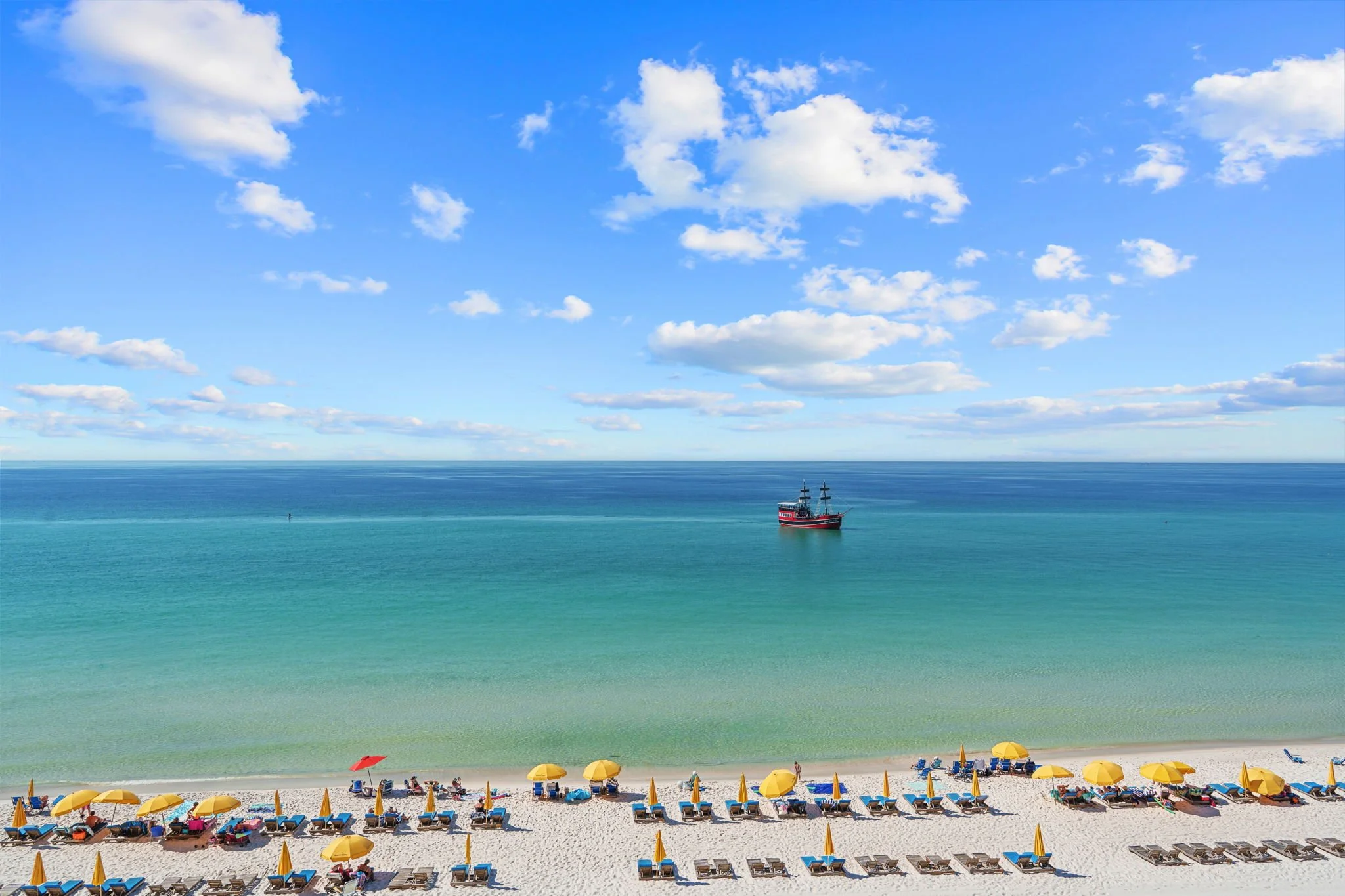 A beach scene with yellow umbrellas and lounge chairs on white sand, clear turquoise water, a boat in the distance, and a partly cloudy blue sky in Panama City Beach