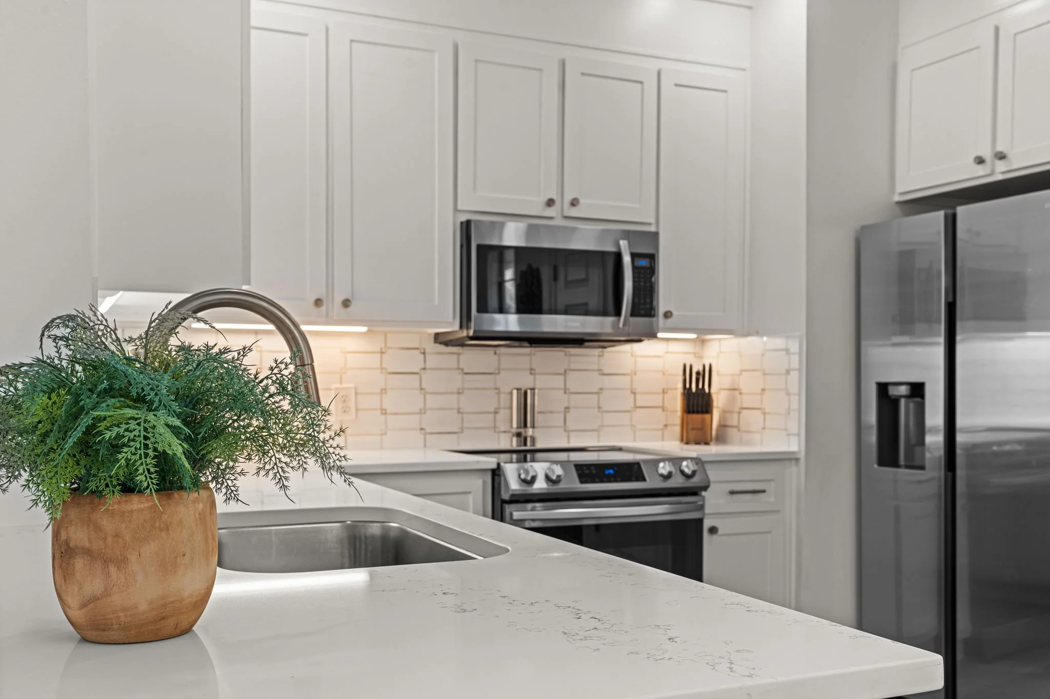 Modern kitchen with white cabinets, stainless steel microwave, oven, and refrigerator. A potted green plant on a white countertop near a stainless steel sink.