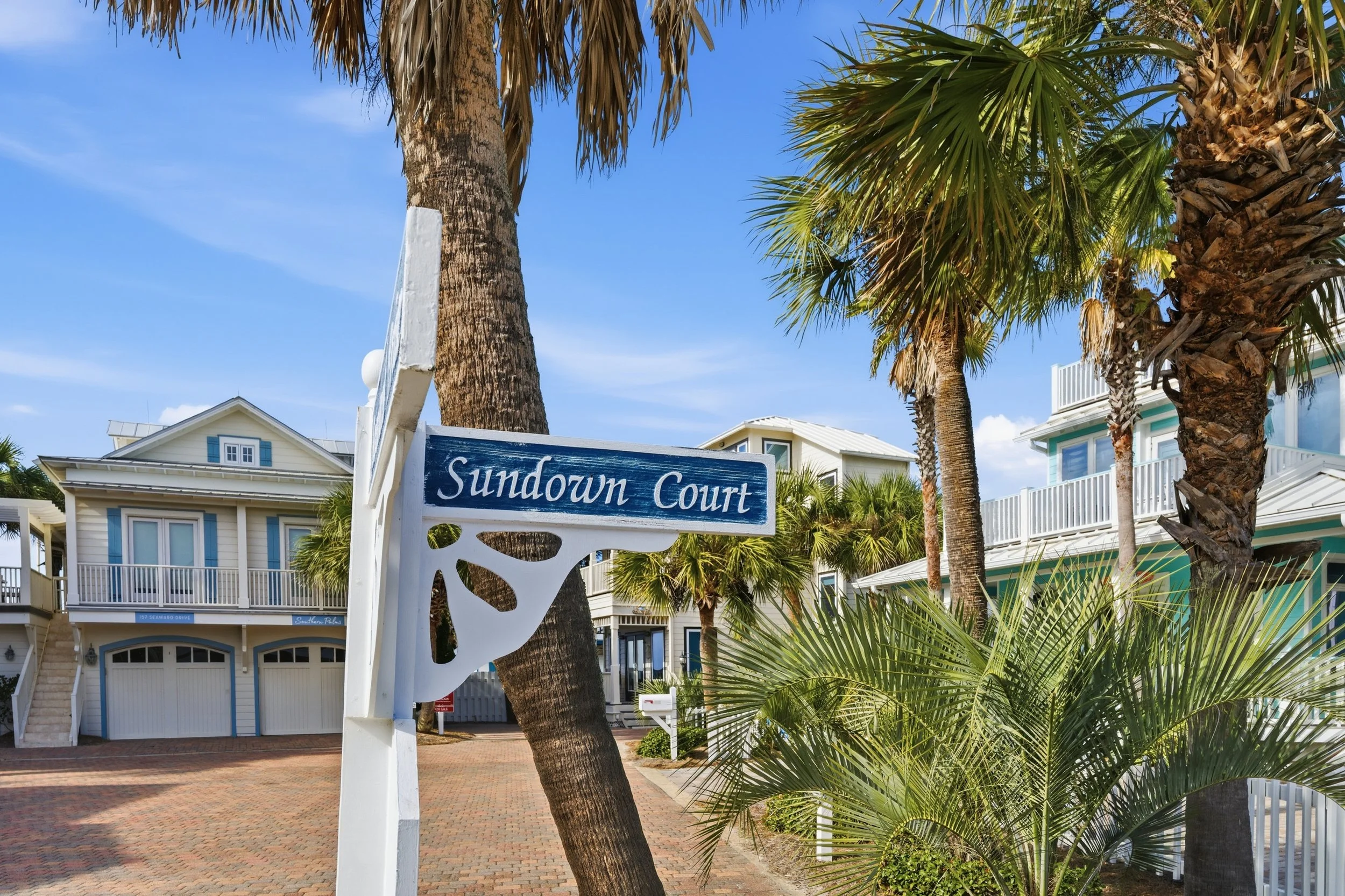 Colorful houses with white railings among palm trees on a sunny day, with a street sign reading 'Sundown Court' in the foreground a image to help sell real estate