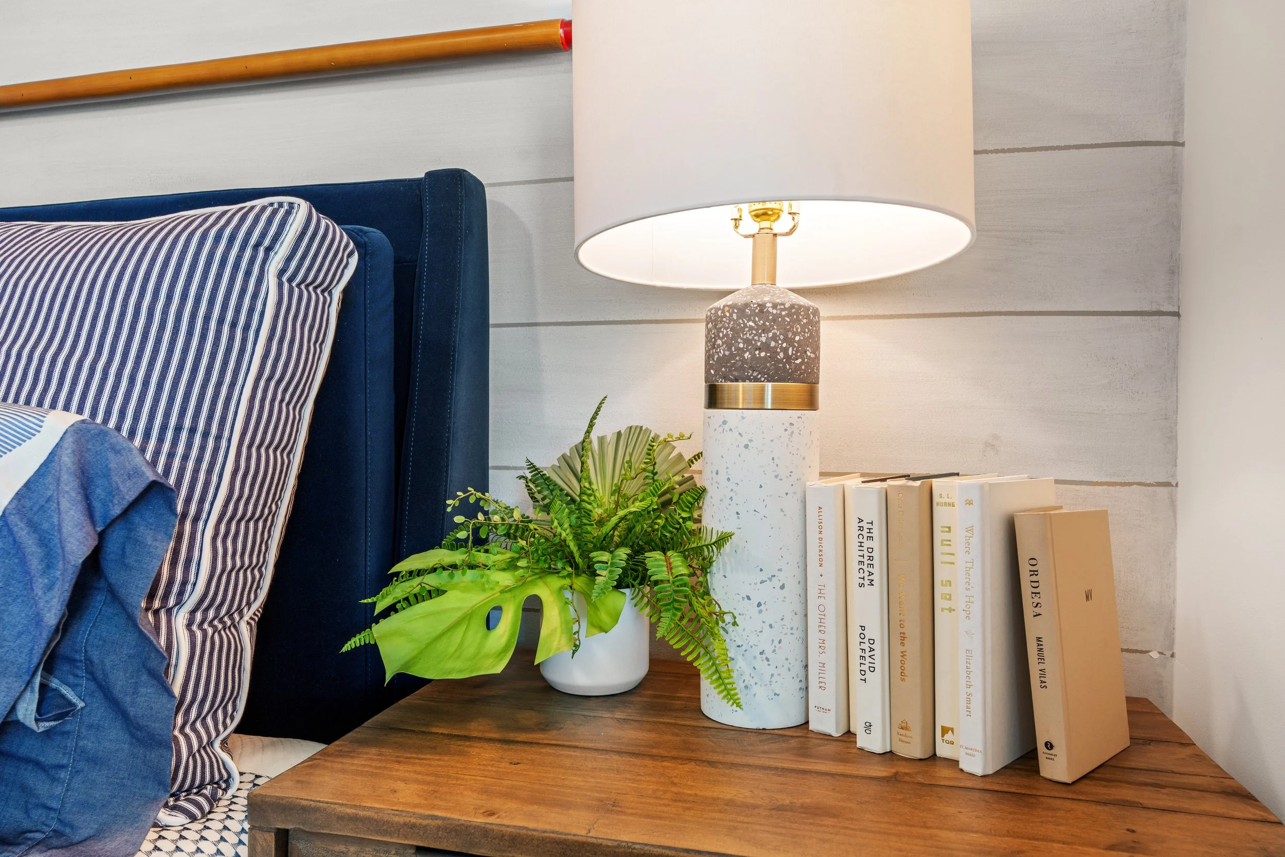 Close-up of a wooden bedside table with a table lamp, a small potted fern, and several books, against a white wall.