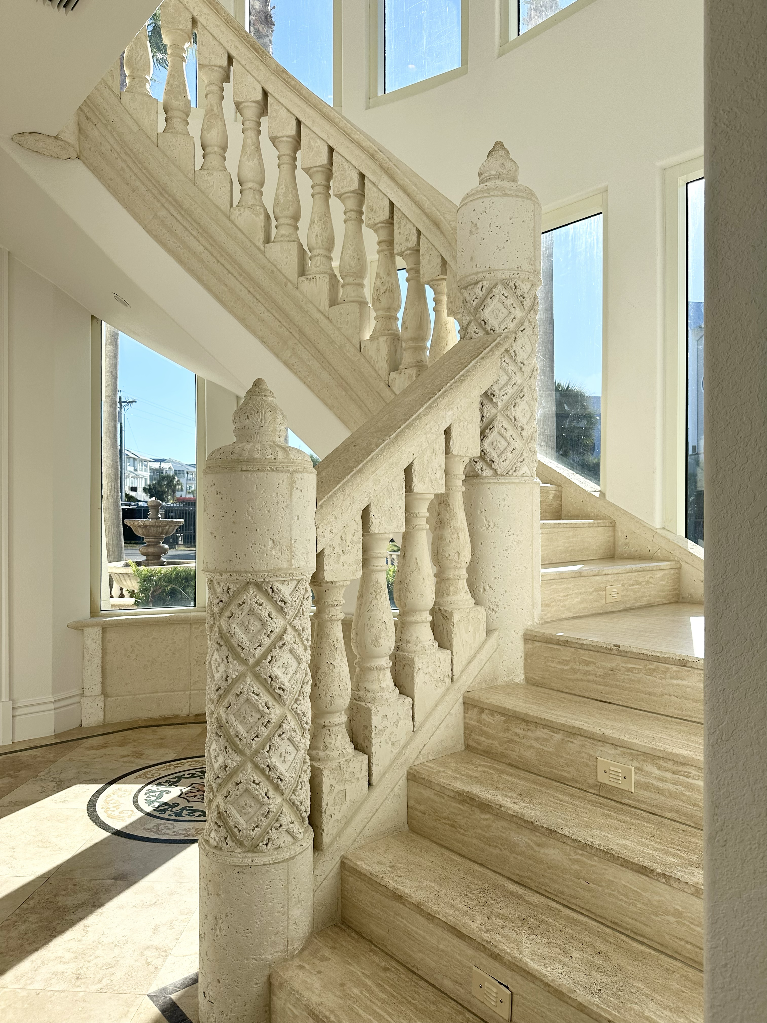 A staircase with ornate carved stone balusters and newel posts, illuminated by large windows allowing natural sunlight to fill the space.
