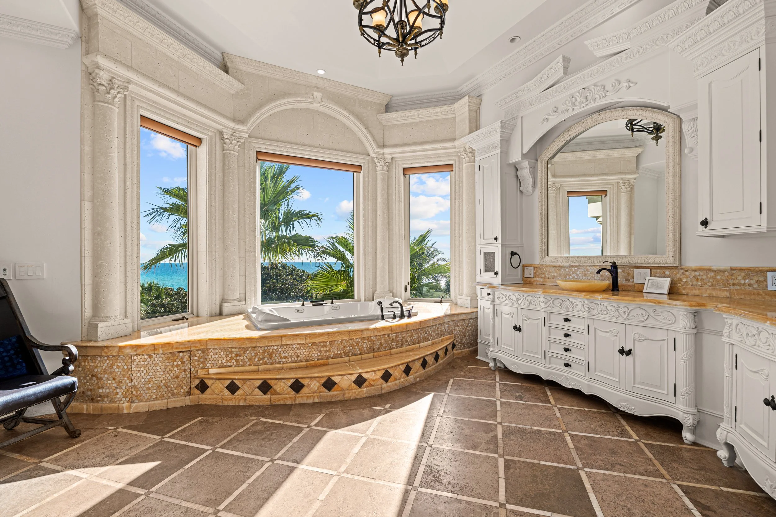 Luxurious bathroom with large windows overlooking palm trees and the ocean, white ornate cabinetry, and a bathtub beneath the windows.