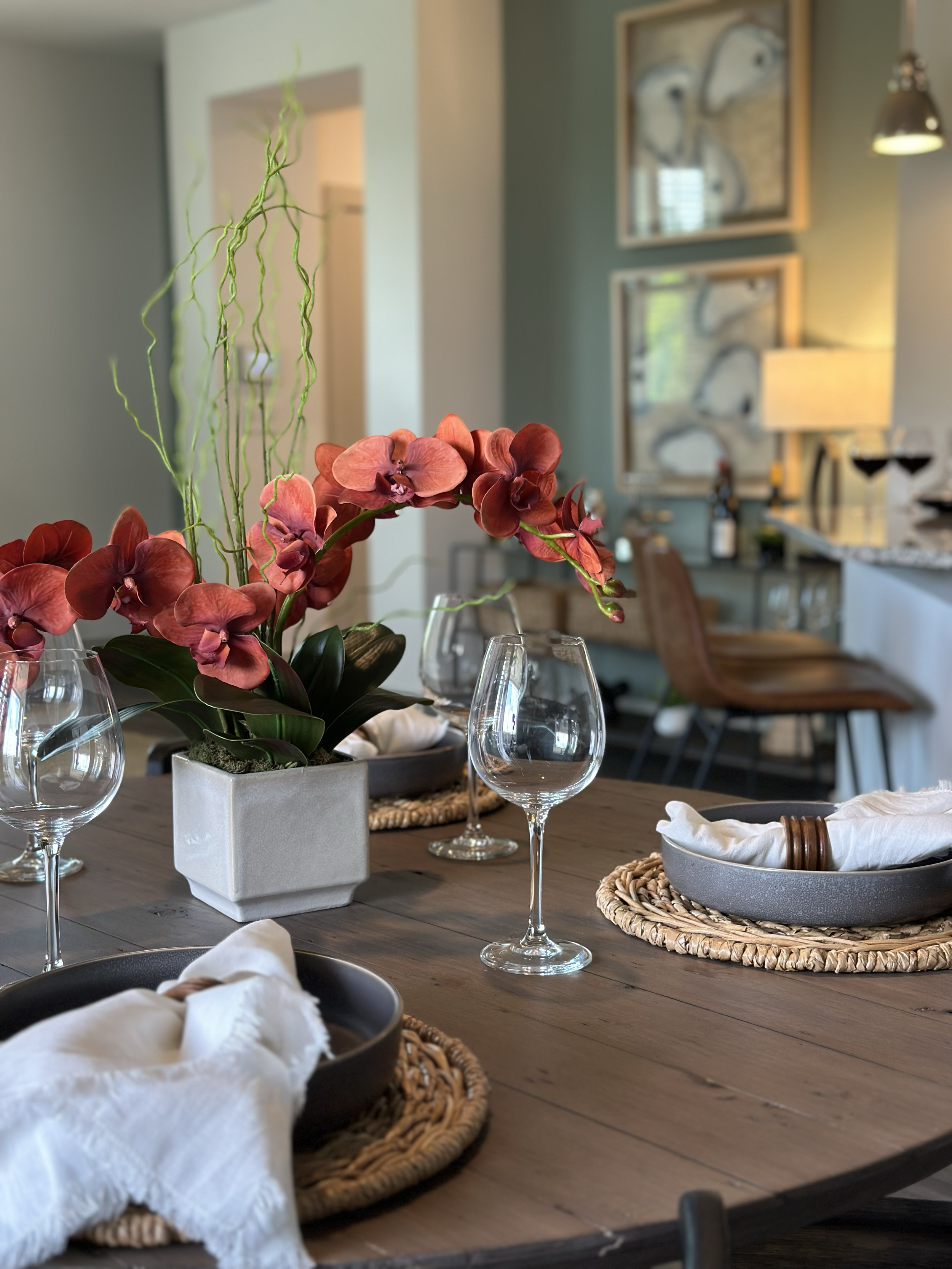 A dining table set with glasses, woven placemats, and napkins, with a centerpiece of pink and red orchids and green curly branches, in a decorated room with artwork and a sideboard in the background. A image that sells "luxury"