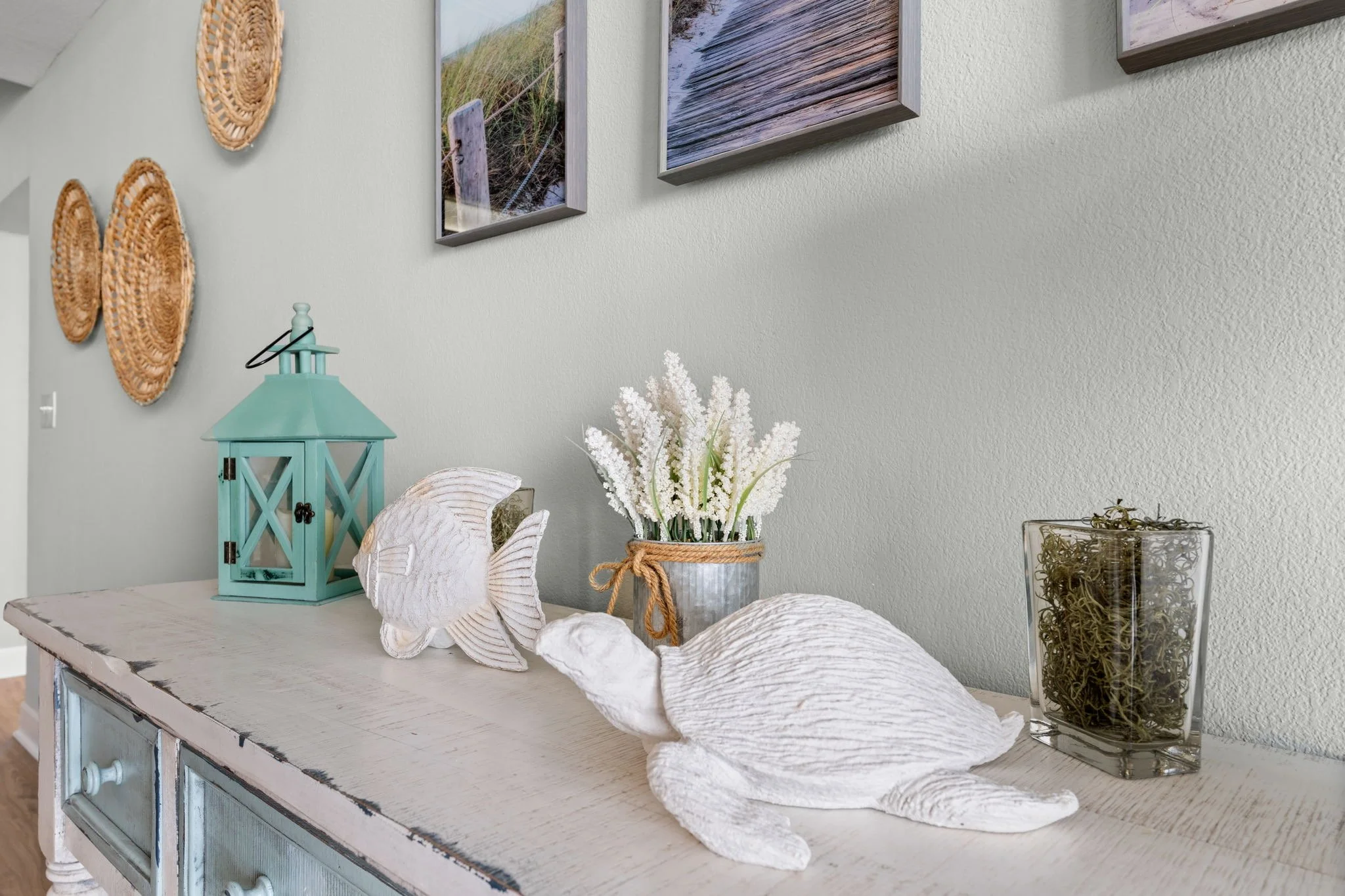 Decorative coastal-themed items on a distressed white sideboard, including white carved turtle and fish sculptures, a glass container with moss, a small potted white flowers, a blue lantern, and wall art depicting beach scenes.