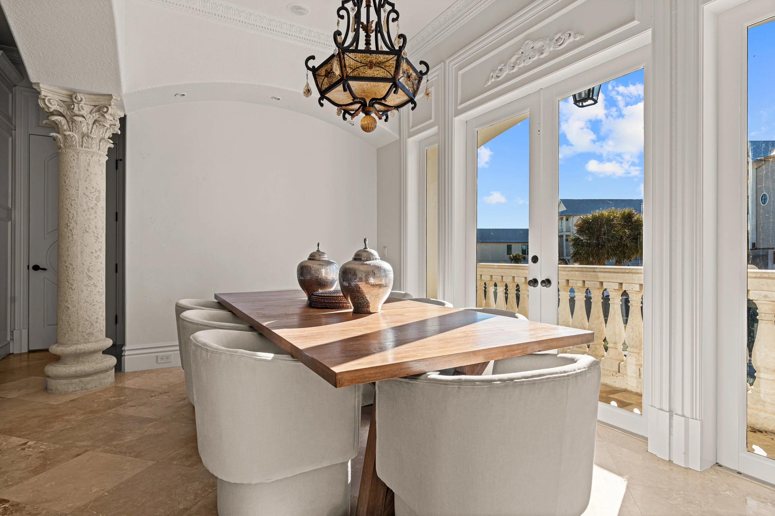 Bright dining room with a wooden table and plush beige chairs, large glass double doors leading to an outdoor balcony with a view of blue sky and trees, ornate chandelier hanging from ceiling, decorative columns, and tiled floor.