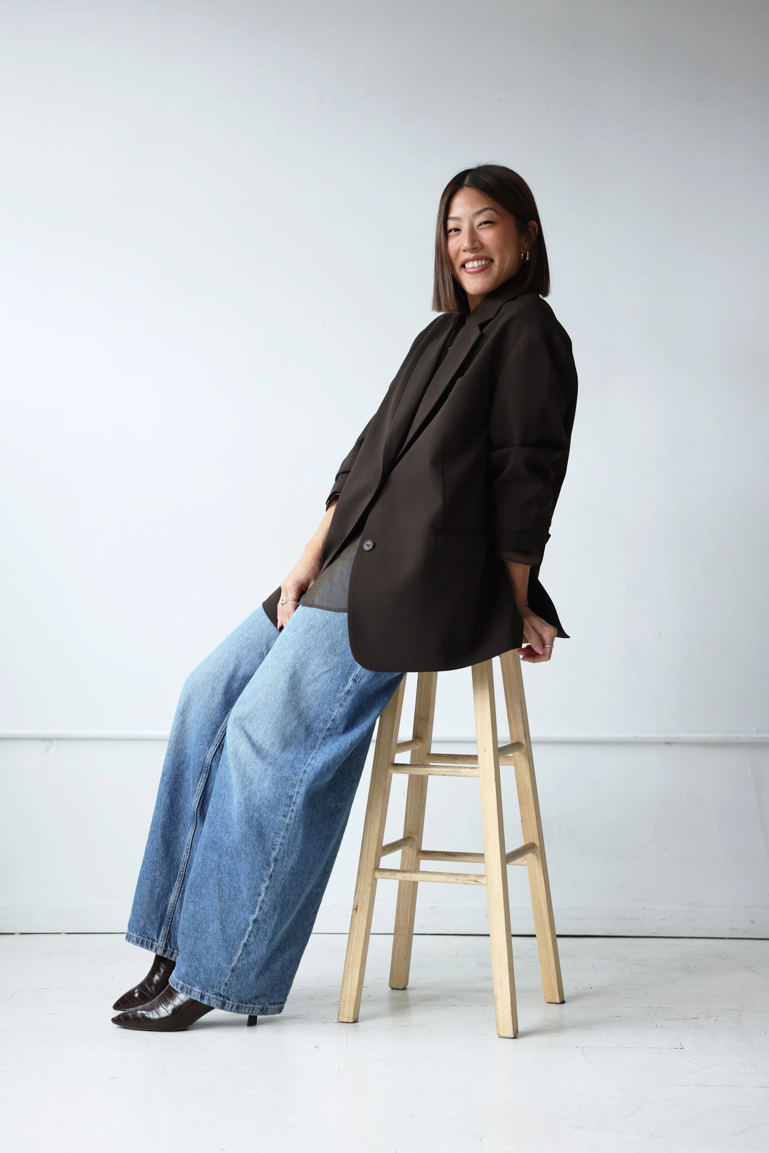 A woman with short, dark hair smiling, sitting on a wooden stool, wearing a brown blazer, wide-leg jeans, and brown boots, posed against a plain white wall.