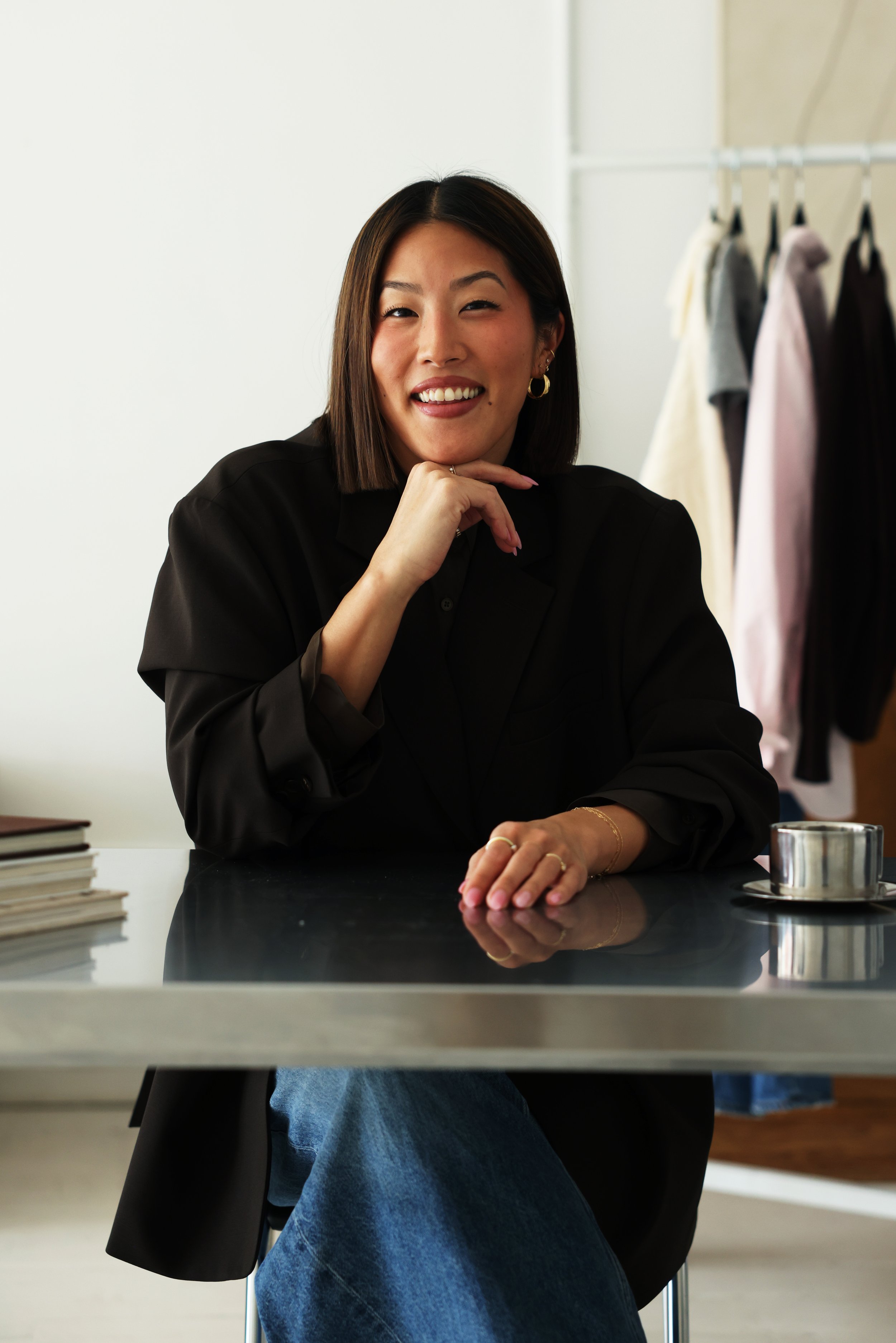 A woman with shoulder-length brown hair smiling at the camera, wearing hoop earrings and a brown blazer, sitting at a chrome table with a stack of books and a metallic cup nearby, in a room with a white background and clothes hanging on a rack.