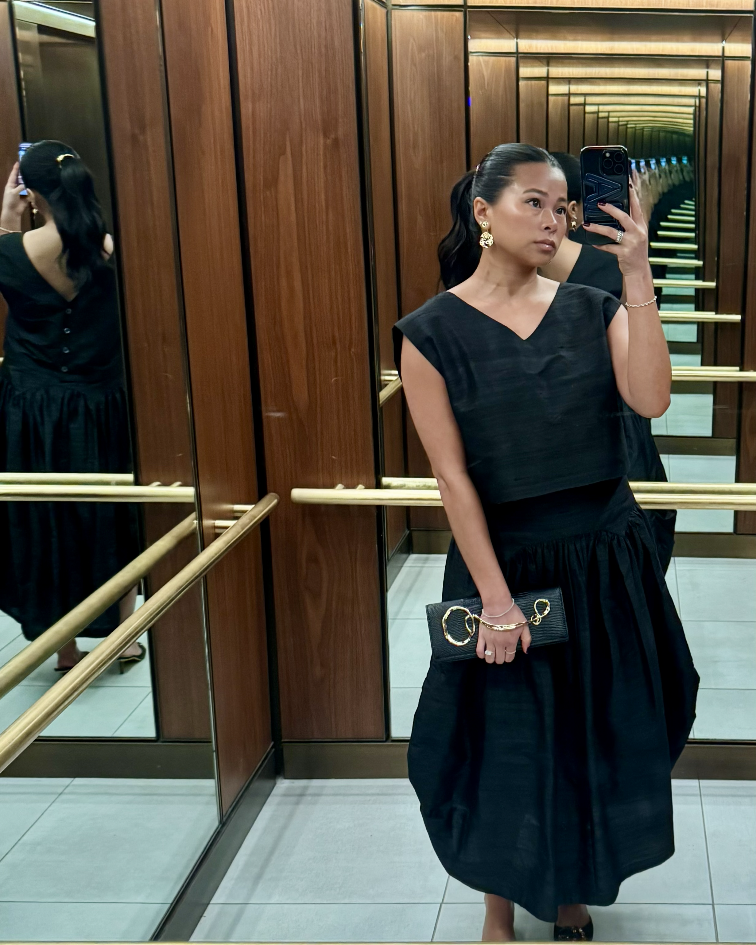 A woman taking a mirror selfie in an elevator with wood-paneled walls. She has dark hair styled in waves, wearing a black dress with cap sleeves, gold earrings, and holding a black clutch with large gold rings. The elevator has reflective surfaces and brass handrails.