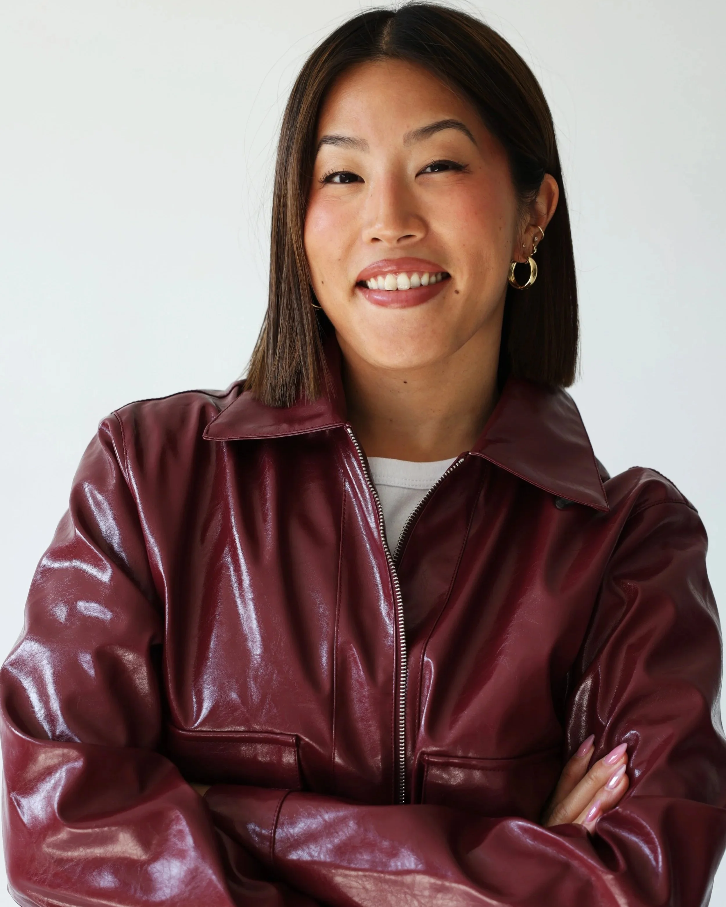 A woman with shoulder-length brown hair, wearing a maroon leather jacket, smiling, standing against a plain white background.