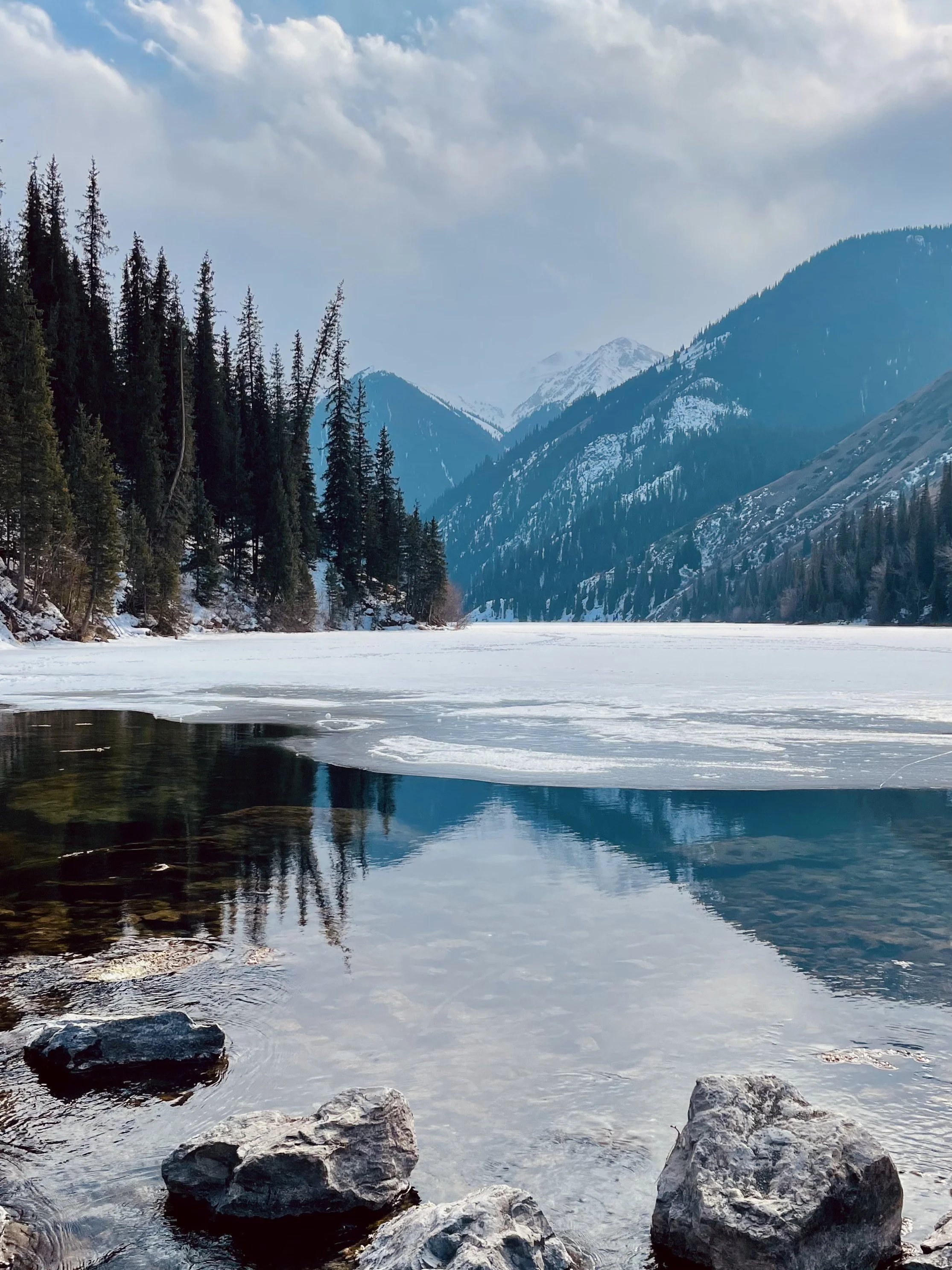 Snow-covered mountains and a partially frozen lake with rocks in the foreground and evergreen trees along the shoreline.