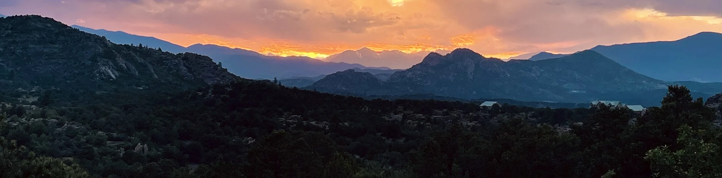 Sunset over mountains with orange and purple sky, silhouette of trees and scattered buildings in the foreground.