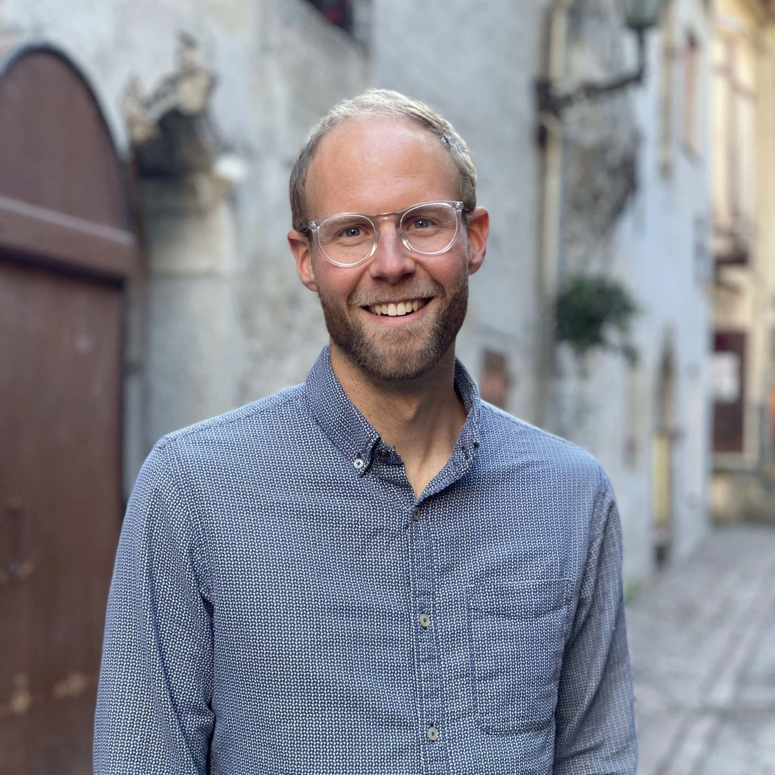 A smiling man with glasses, beard, and short hair, wearing a patterned button-up shirt, standing outdoors on a cobblestone street with stone and stucco buildings in the background.