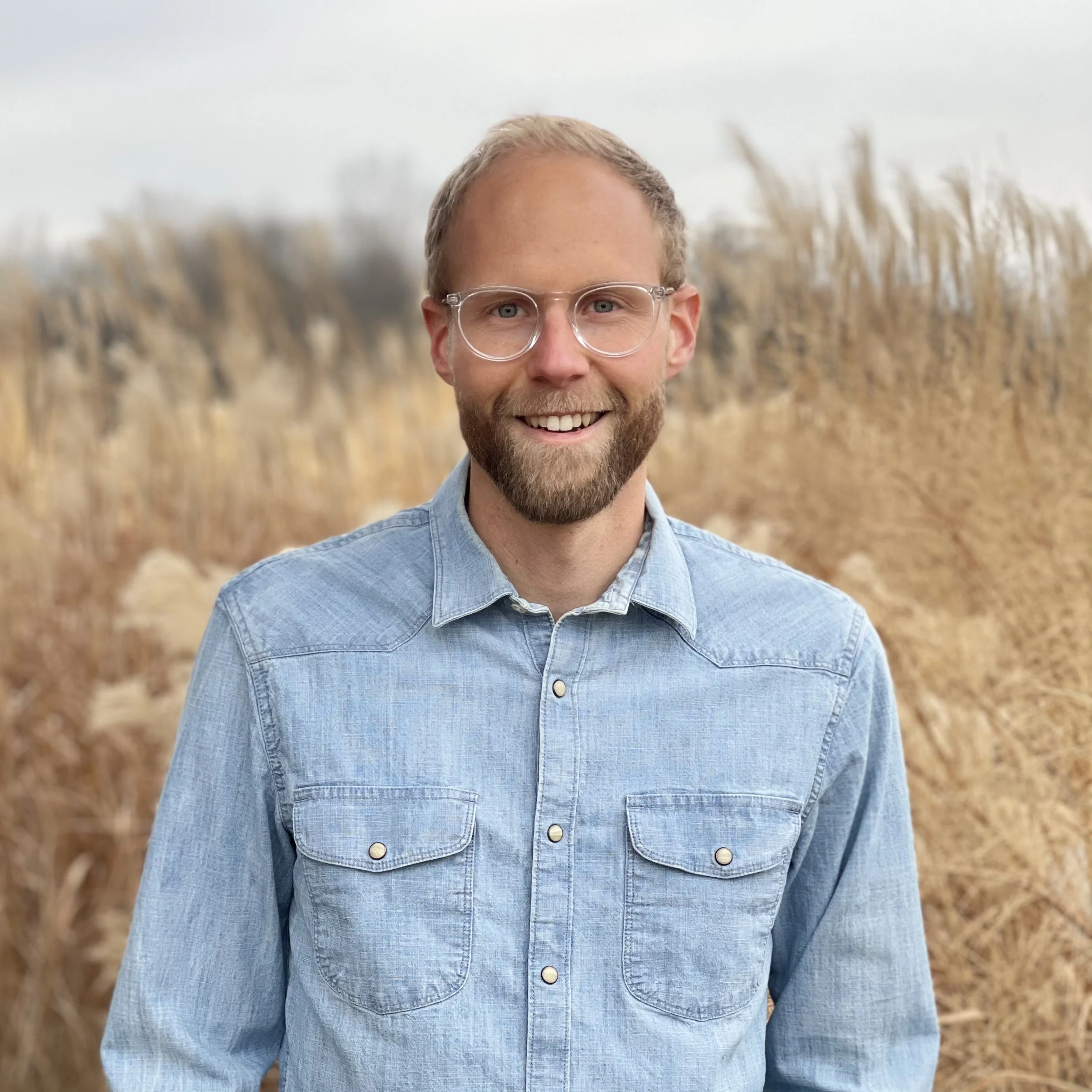 A smiling man with glasses and a beard standing outdoors in front of tall brown grass on a cloudy day.