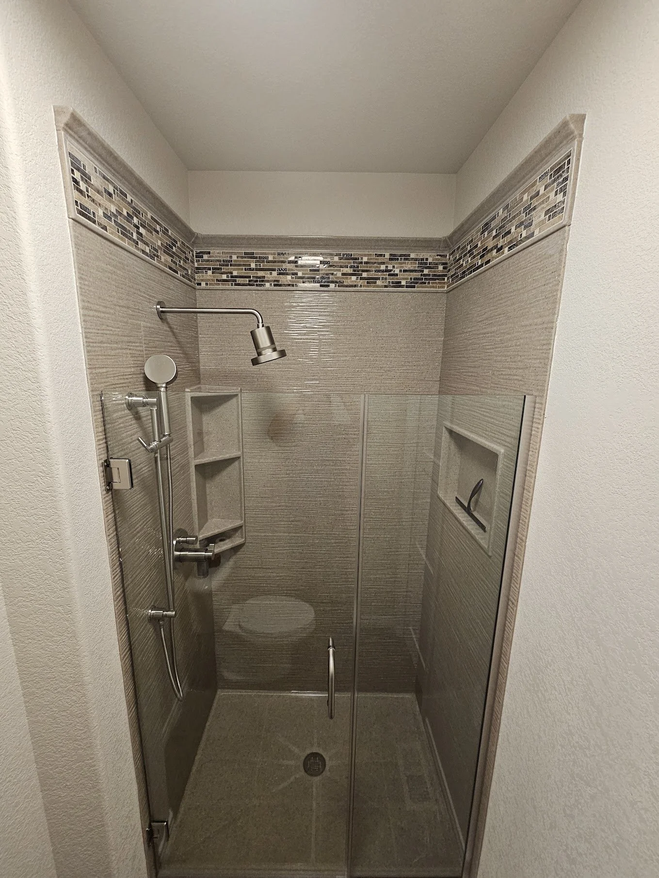Inside a small tiled shower with a glass door, built-in shelving, a wall-mounted showerhead, and a small niche for toiletries.