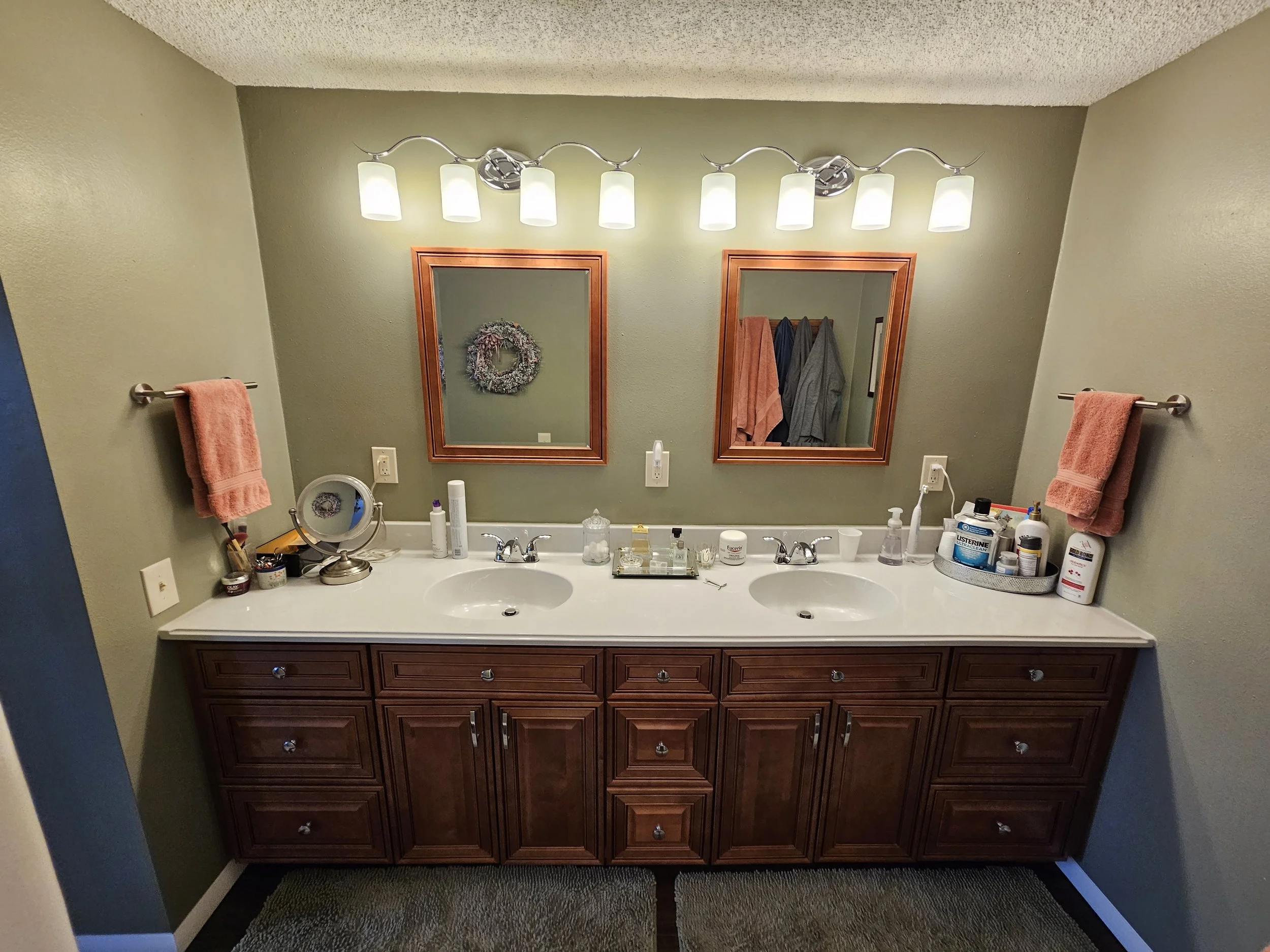Bathroom vanity with two sinks, two mirrors with wooden frames, and two wall-mounted light fixtures above the mirrors. Pink hand towels on towel bars, various toiletries on the countertop, and a green wall background.