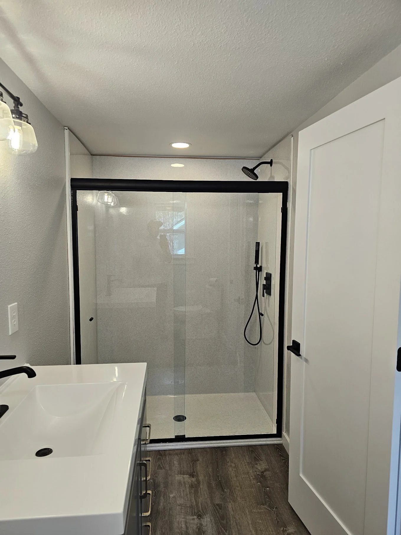 Modern bathroom with a glass shower enclosure, black frame, showerhead, and handheld shower; white vanity with black faucet; white walls and door; wood-look flooring.