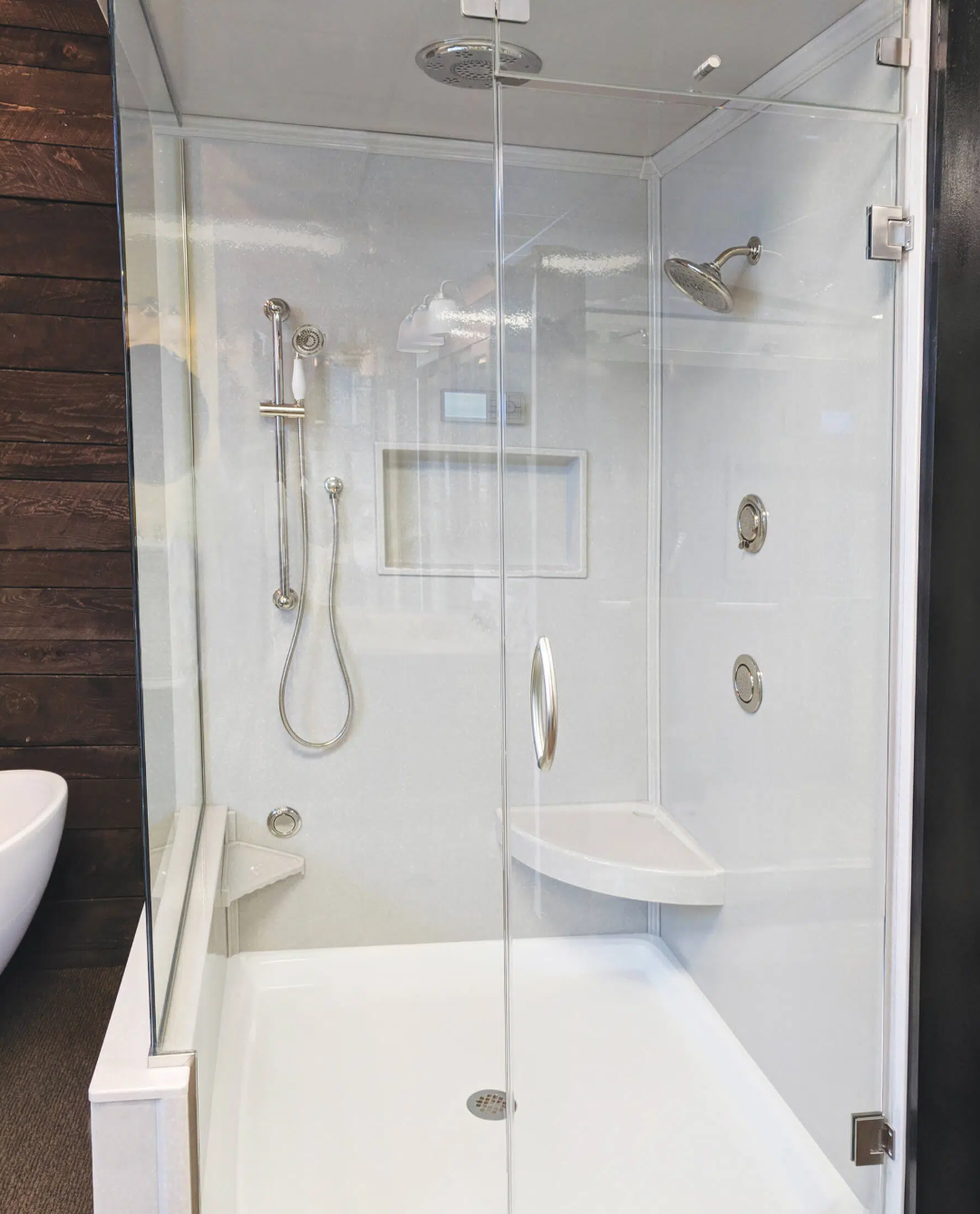 A modern glass shower enclosure with a handheld showerhead, a wall-mounted showerhead, and a built-in corner seat. The shower has white walls and a drain on the floor.