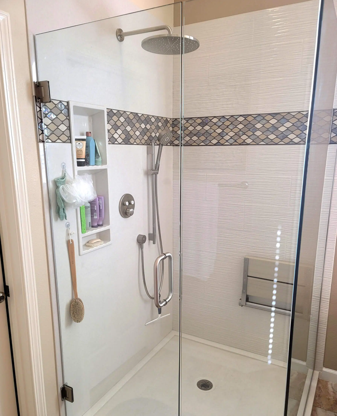 A sitting shower with clear glass walls, built-in shelves with toiletries, a rain showerhead, hand-held shower with adjustable height, and a foldable seat inside a bathroom.