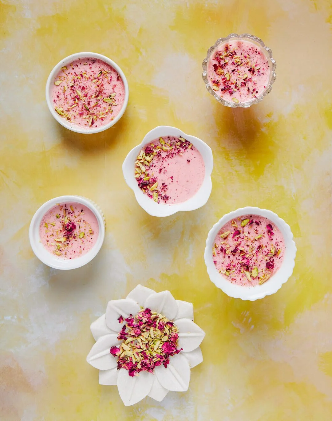 Various bowls and glasses of pink dessert garnished with chopped pistachios and rose petals on a yellow background.