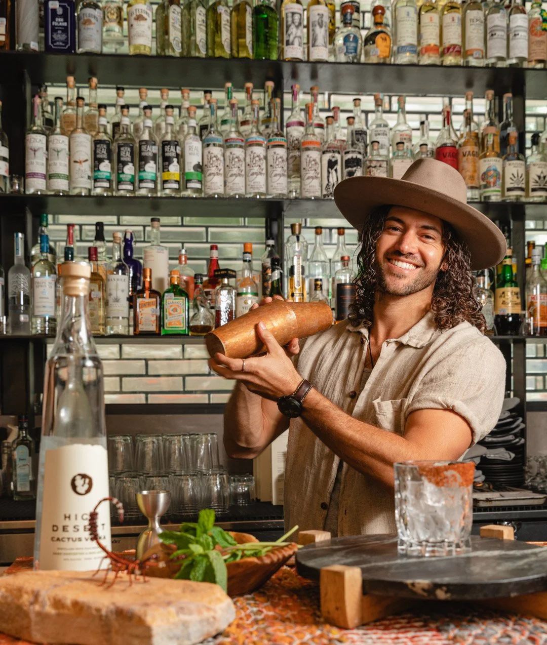 A bartender smiling and holding a copper cocktail shaker in a bar with shelves of liquor bottles in the background.