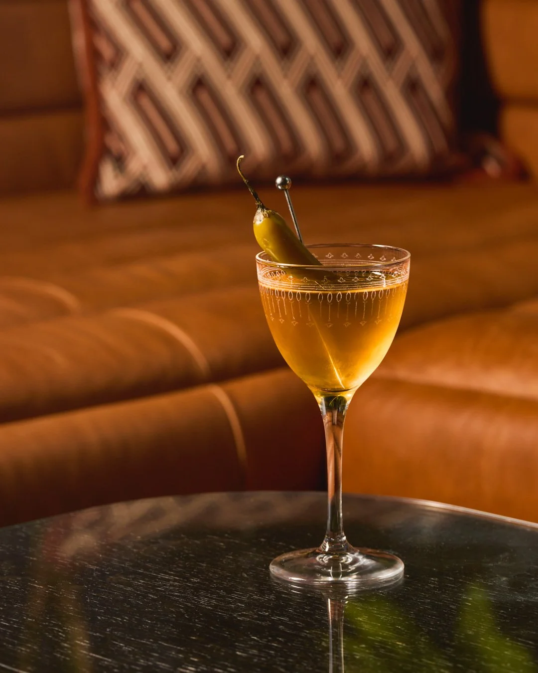 A cocktail glass with an orange-colored drink garnished with a green chili pepper pierced by a cocktail pick, sitting on a dark wooden table with a blurred couch and patterned pillow in the background.