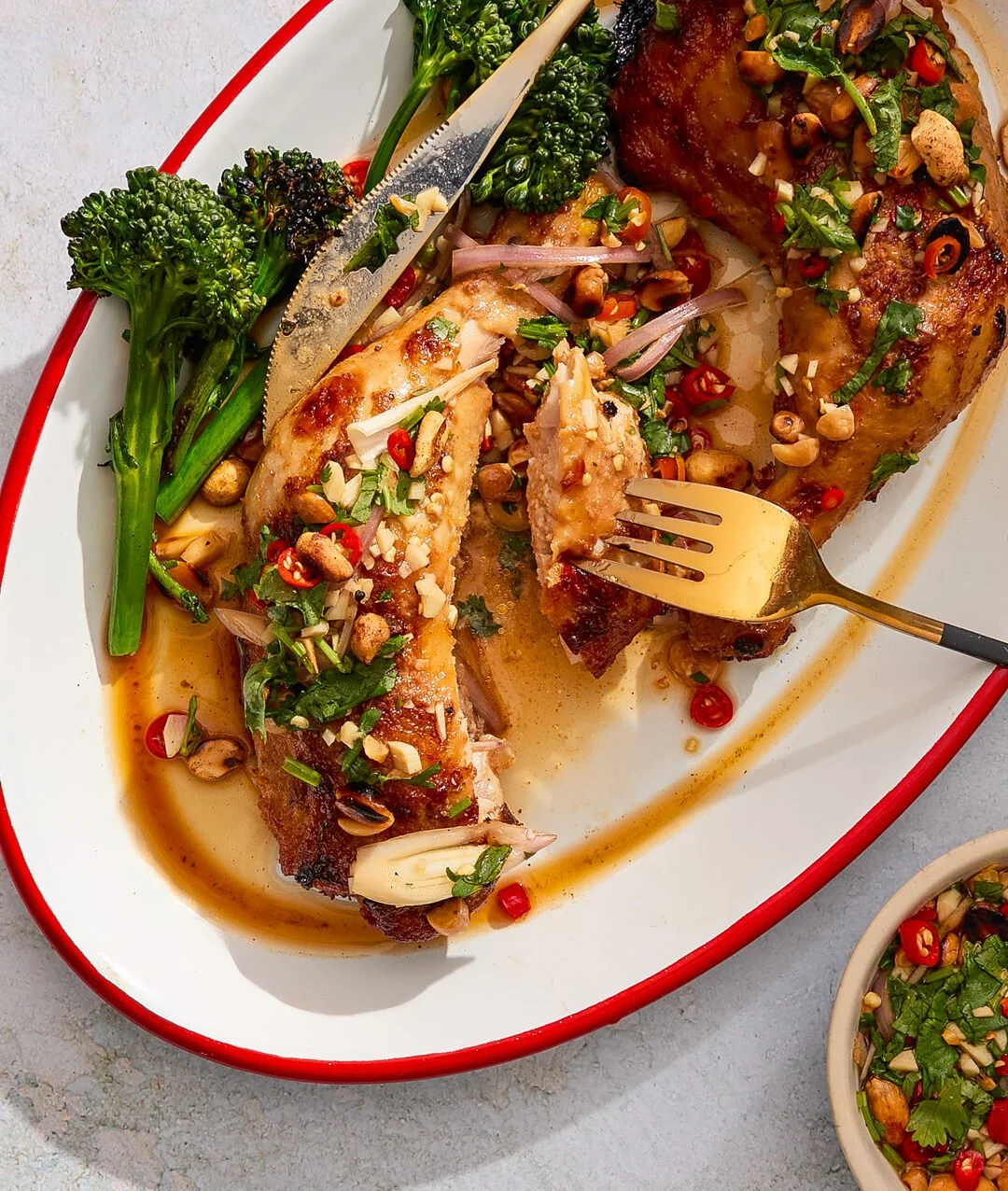 Roasted chicken leg garnished with chopped herbs, sliced red chili, garlic, and crushed nuts, served with broccoli and a side of sauce.
