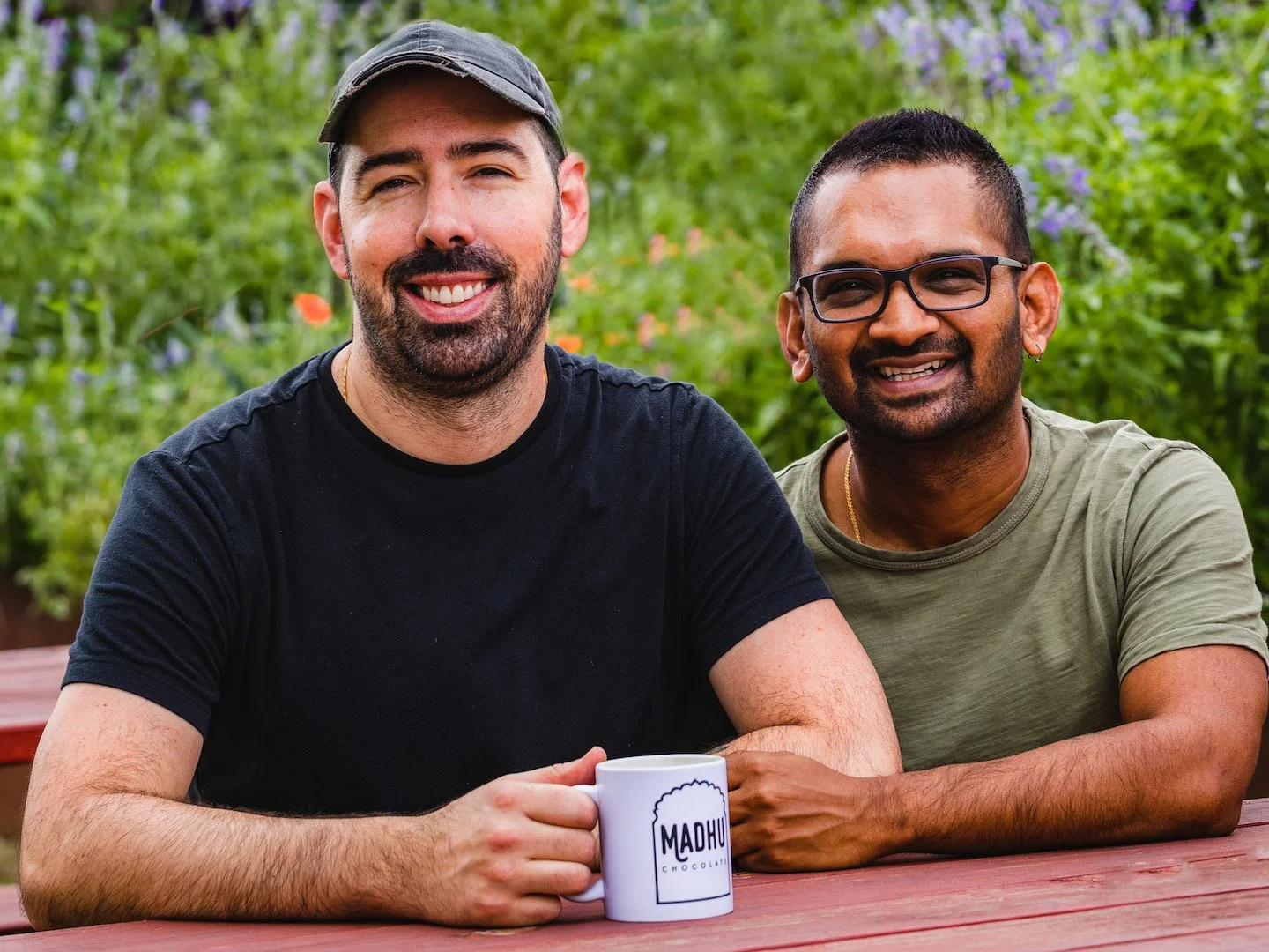 Two men smiling and sitting outdoors at a wooden table, one holding a mug with "MADHU" written on it, lush green background with flowers.