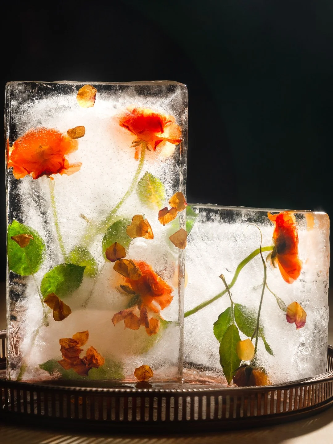Ice sculptures with embedded orange and yellow flowers and green leaves, set on a tray against a dark background.