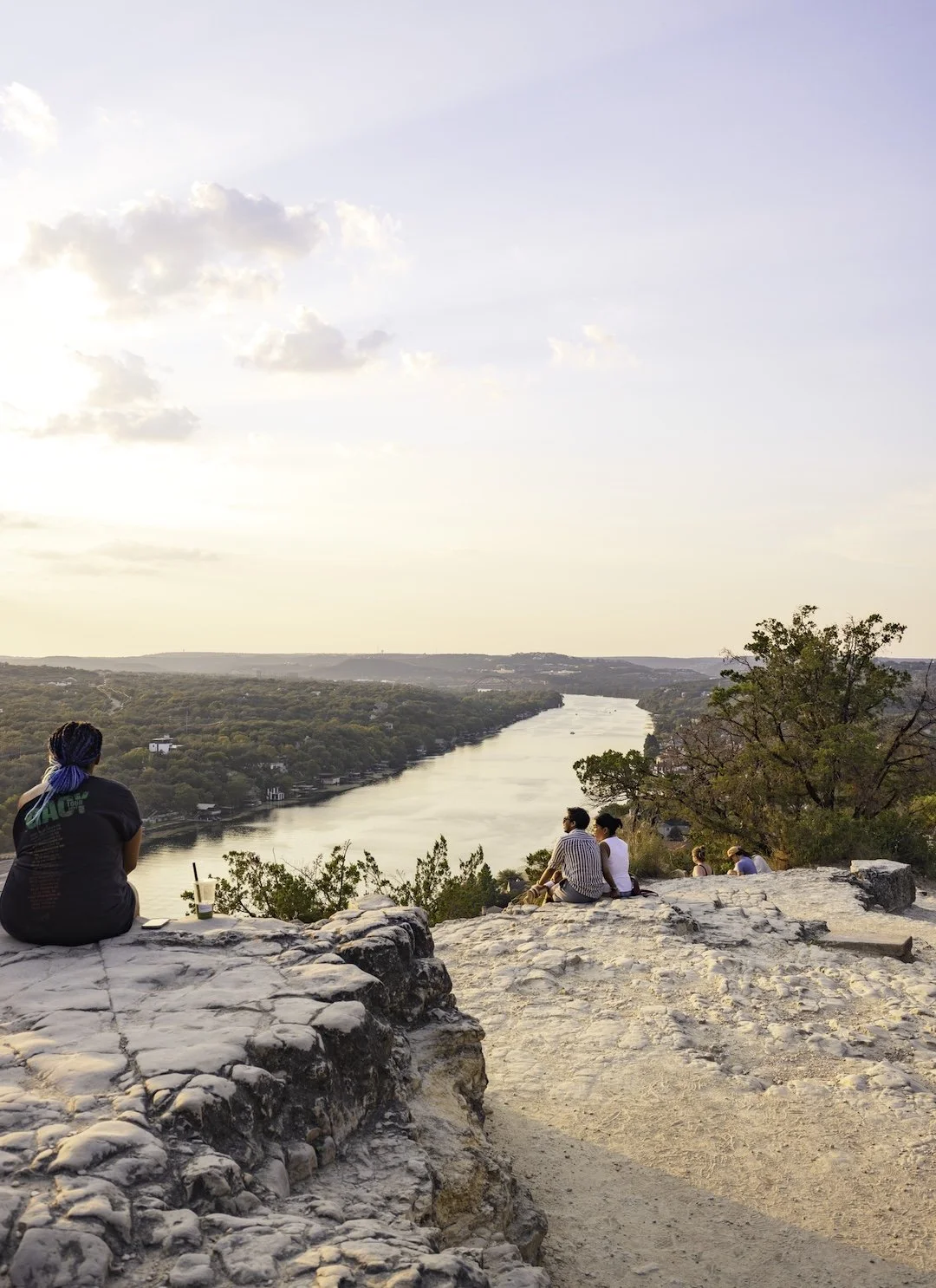 People sitting on rocky ledge overlooking a river at sunset with trees on the banks and a cloudy sky.