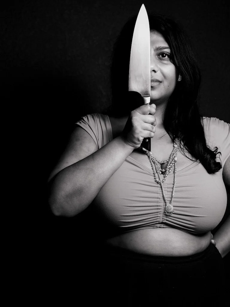 A woman with dark hair holding a large kitchen knife in front of her face, half of her face visible, against a dark background.