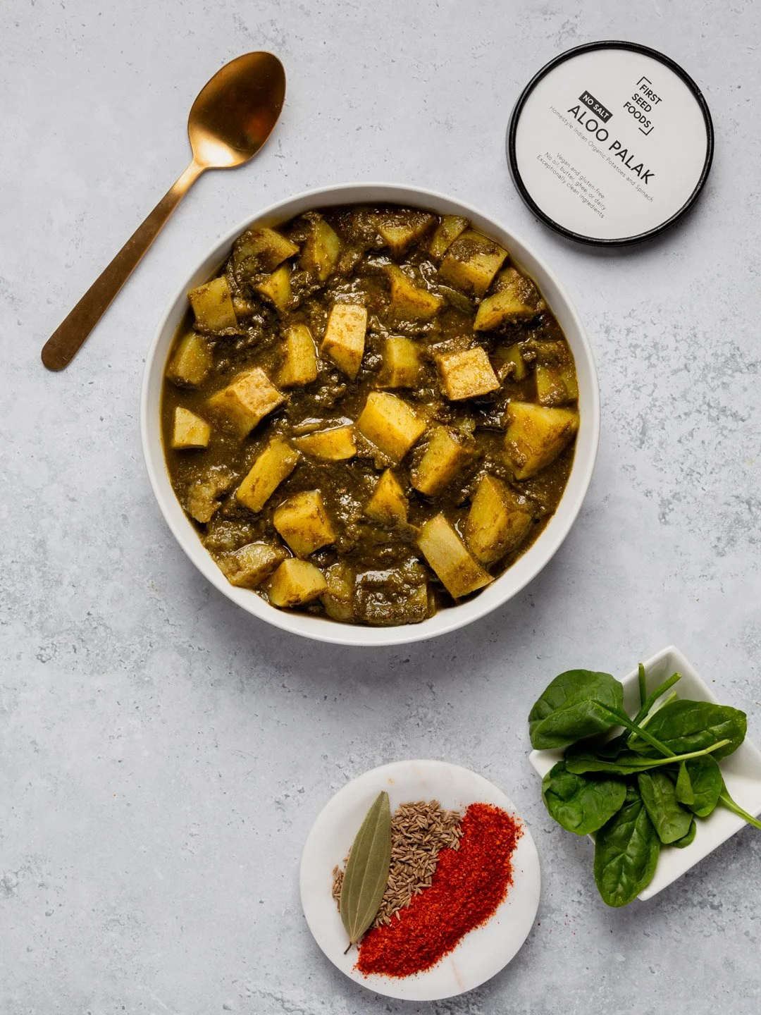 A bowl of Indian-style eggplant curry with garlic and spices, served on a white surface with a gold spoon, a container of 'Aloo Palak' in the background, and a small plate of cumin seeds, red chili powder, and a bay leaf, along with a small bowl of f