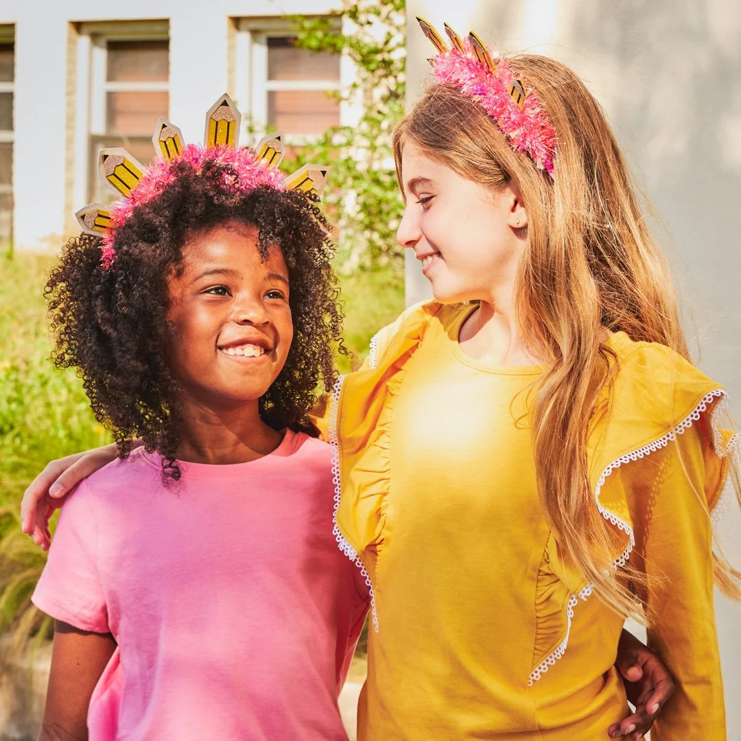 Two young girls wearing colorful crowns with pink feathers, smiling and looking at each other outdoors.