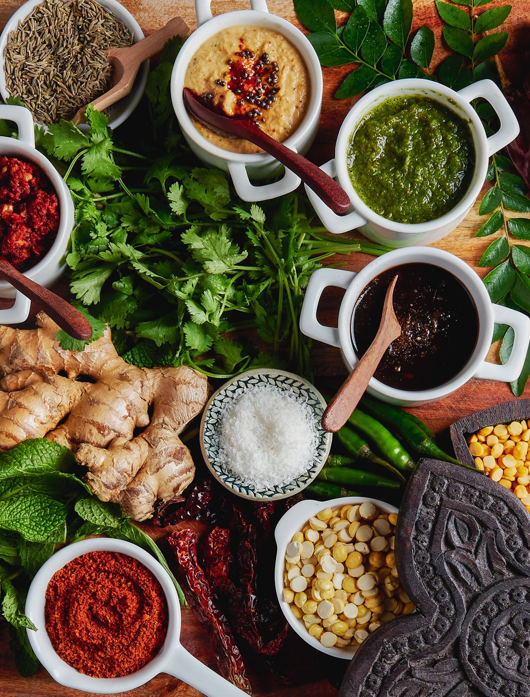 Assorted spices, herbs, and sauces for cooking on a wooden surface, including ginger, dried chili peppers, cilantro, cumin seeds, coconut flakes, and sauces in small white bowls with wooden spoons.