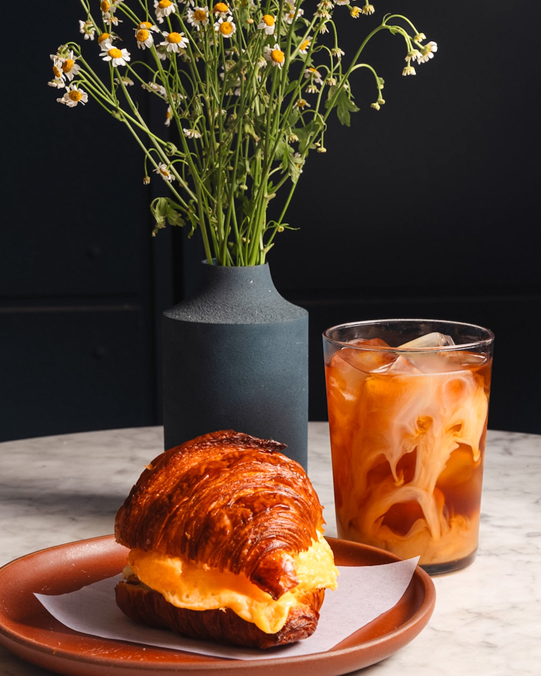 A croissant sandwich with egg and cheese on a terracotta plate in front of a glass of iced coffee with cream swirls, and a vase of small white daisies on a light-colored marble surface against a black background.