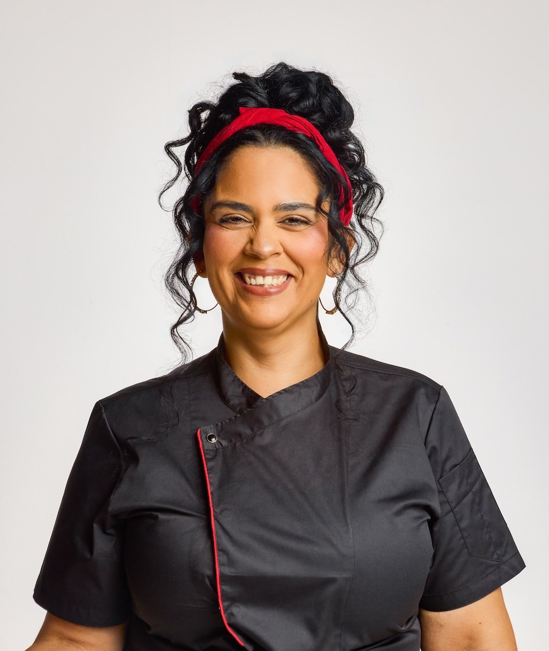 Smiling woman with curly black hair, red headband, wearing black chef's jacket, against a plain white background.