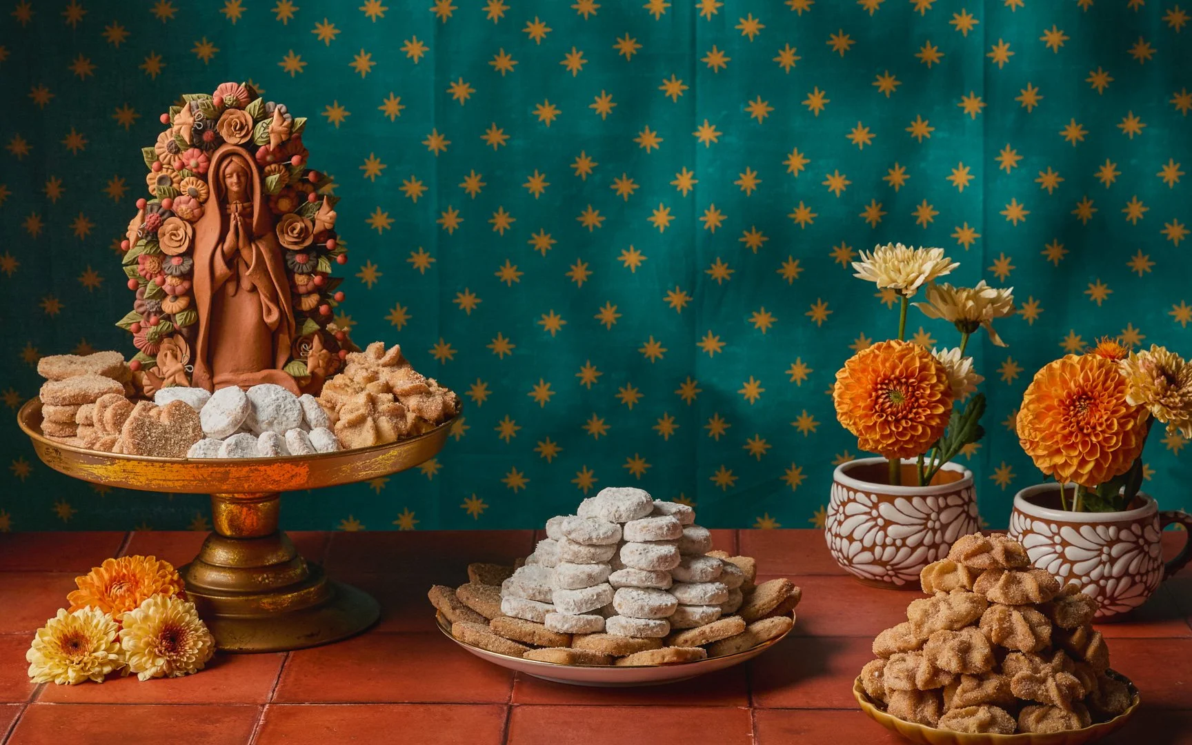 A festive altar with a statue of Our Lady surrounded by flowers and traditional Mexican sugar cookies and confections on plates, with a teal backdrop decorated with gold stars.