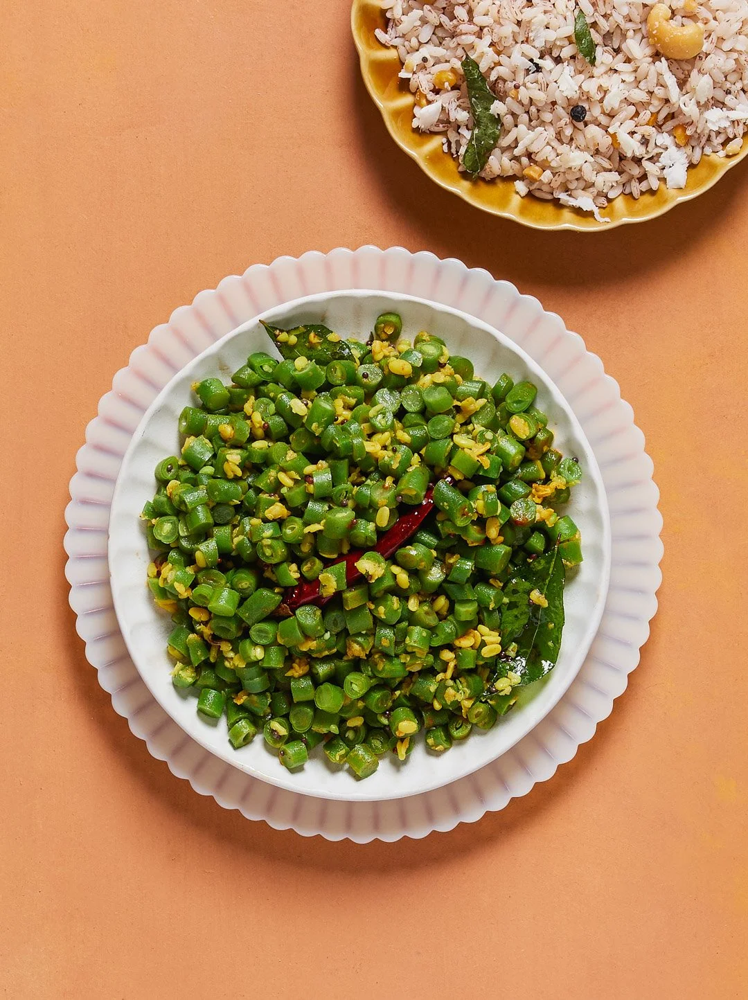 A bowl of chopped green beans garnished with spices and leaves, with a yellow plate of rice in the background.