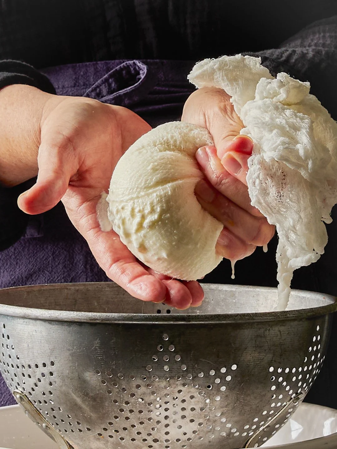 Person squeezing fresh curd cheese with their hands over a metal strainer.