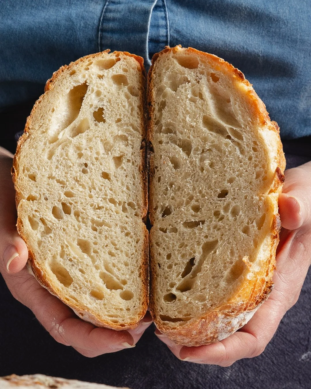 Hands holding a halved loaf of rustic bread with a golden crust and airy, open crumb structure.