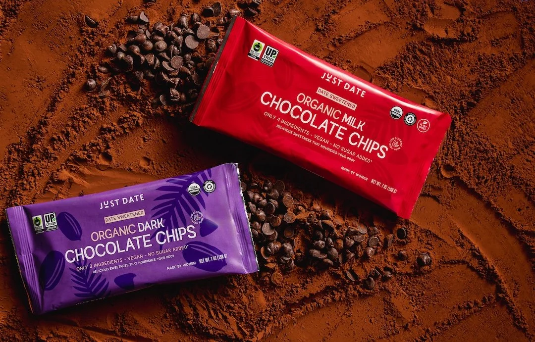 Two packages of organic chocolate chips, one dark and one milk, placed on a surface sprinkled with cocoa powder, with some chocolate chips scattered around.