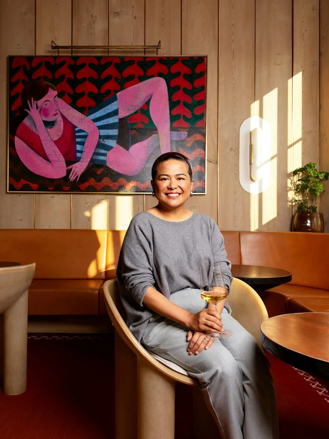 A woman sitting in a modern lounge area, smiling and holding a glass of white wine with sunlight streaming in behind her. There is a colorful abstract painting and a potted plant on a wooden wall.