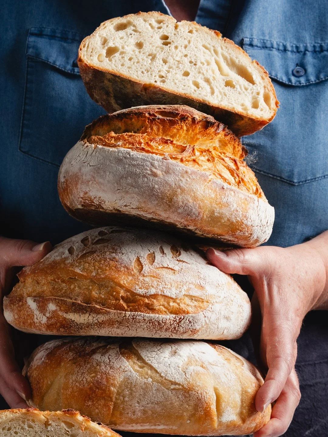 Stack of five freshly baked sourdough bread loaves, with one halved to show the airy interior, held by a person wearing a blue jean shirt.