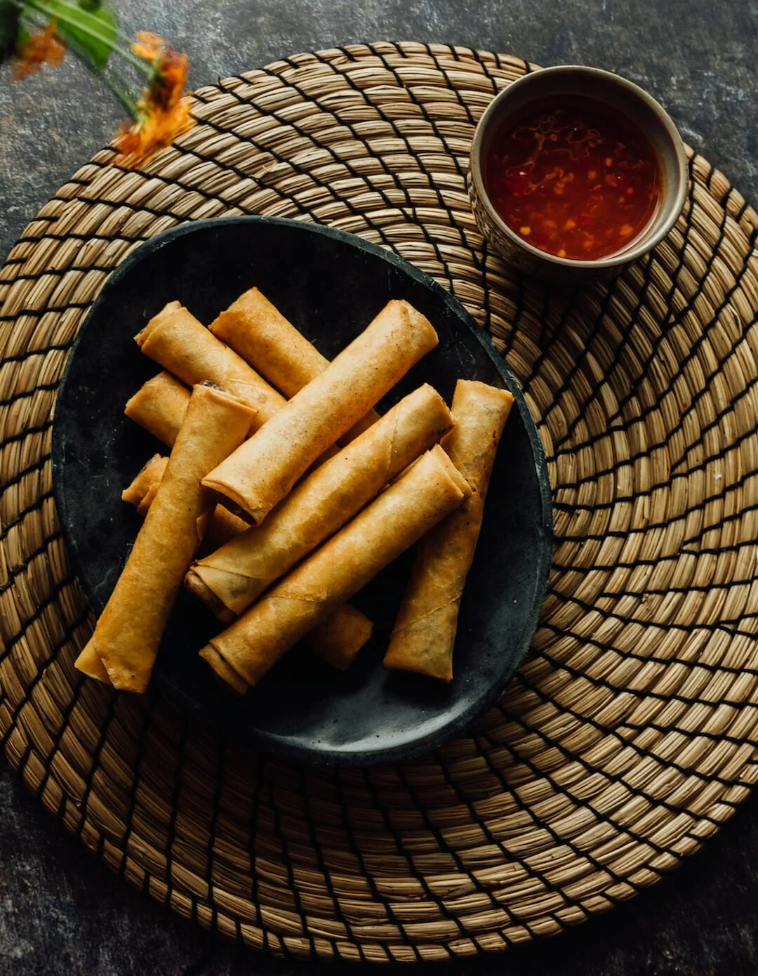A black plate with golden spring rolls on a woven rattan placemat, accompanied by a small bowl of dipping sauce.