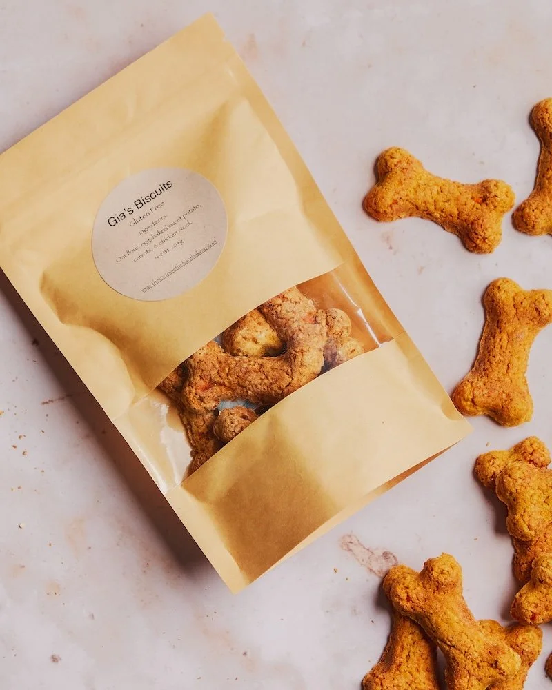 A yellow bag of Gia's Biscuits with a clear window showing bone-shaped dog treats, some of the treats are spilled out on a white surface.