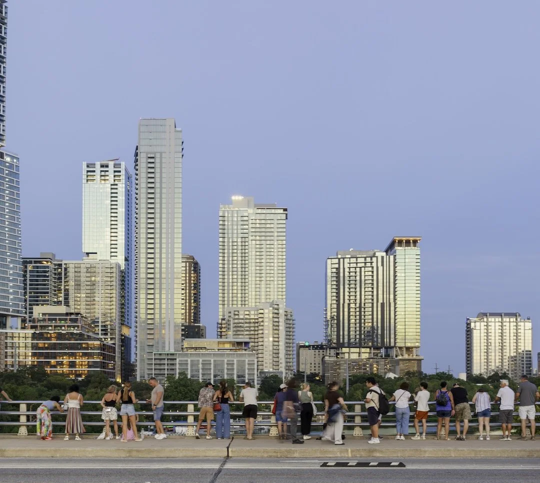 People gathered on a bridge overlooking a city skyline with tall modern buildings during sunset or early evening.