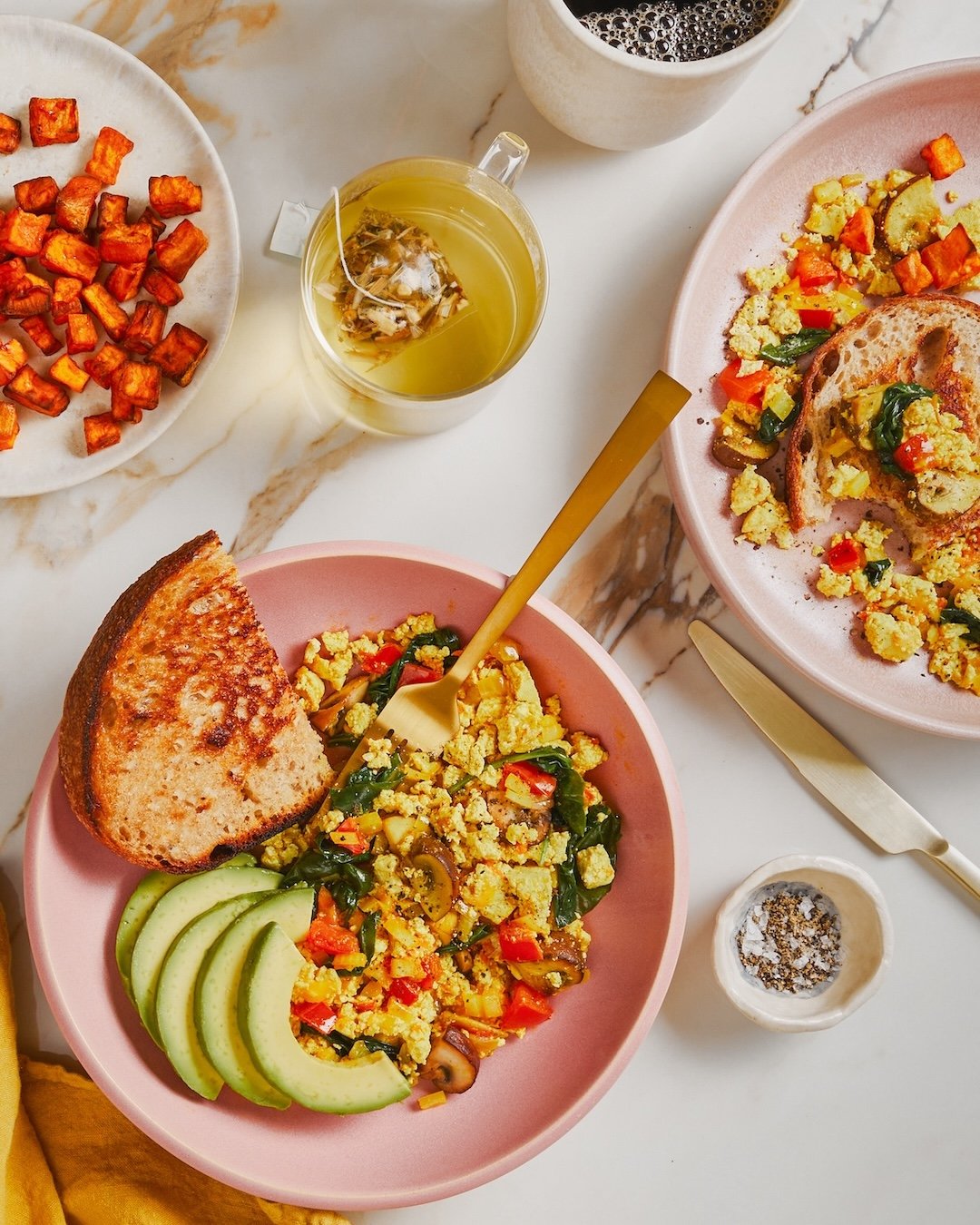 Breakfast spread with scrambled eggs and vegetables on pink plates, alongside sliced avocado and toast, accompanied by roasted sweet potatoes, a cup of coffee, iced tea with tea bag, and black pepper in a small white dish.