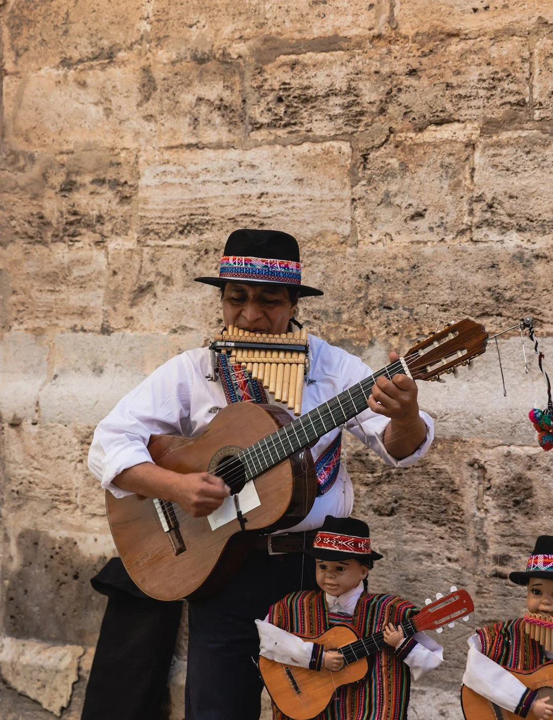 A musician wearing traditional clothing and a wide-brimmed hat, playing guitar and a pan flute, with two dolls dressed in traditional attire, also holding guitars, against a stone wall background.