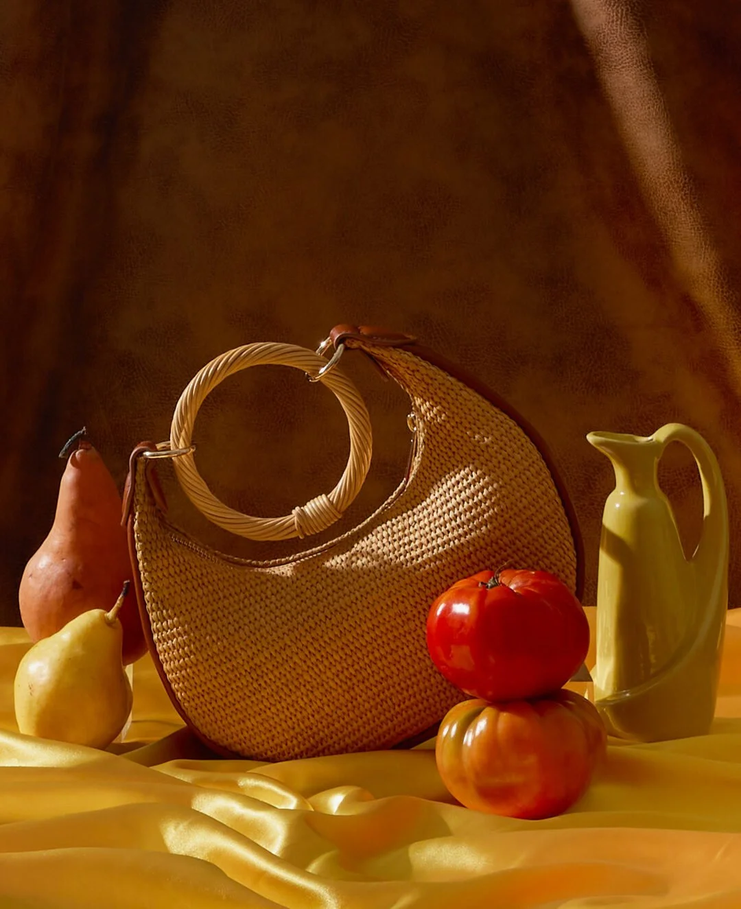 A still life arrangement of a wicker handbag, two pears, two tomatoes stacked, a yellow pitcher, and a yellow satin cloth in the background.