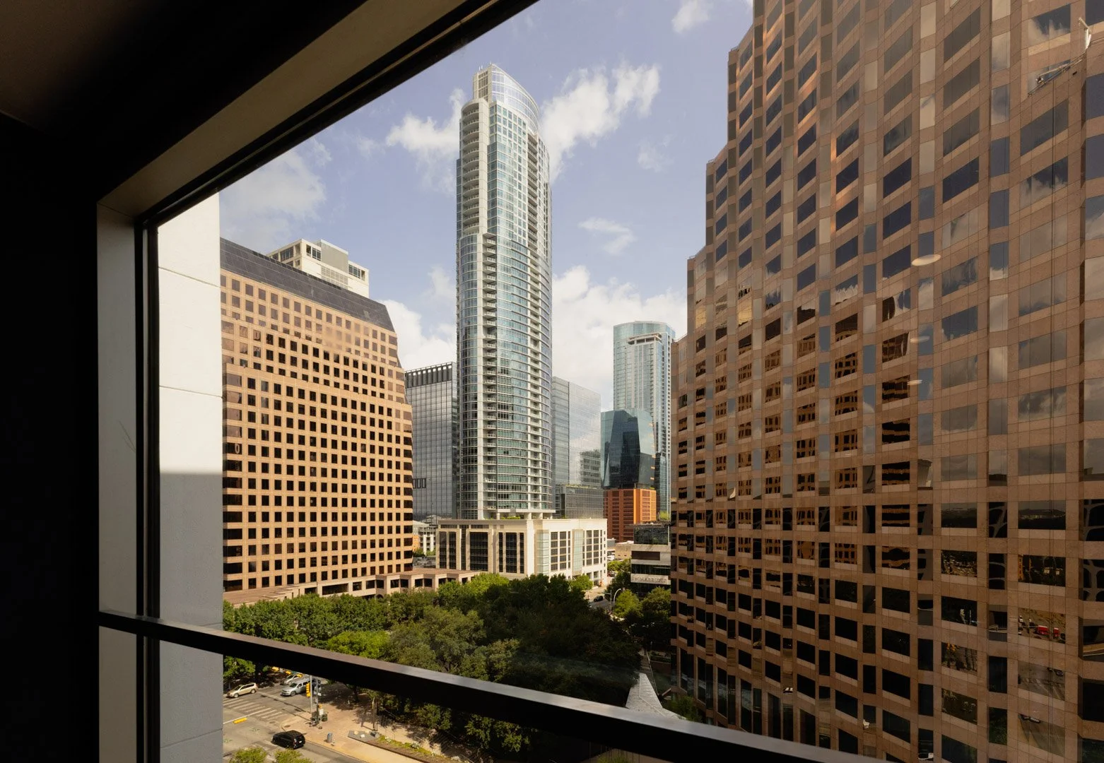 Cityscape view through a window showing tall modern skyscrapers, a partly cloudy sky, and some trees and streets below.