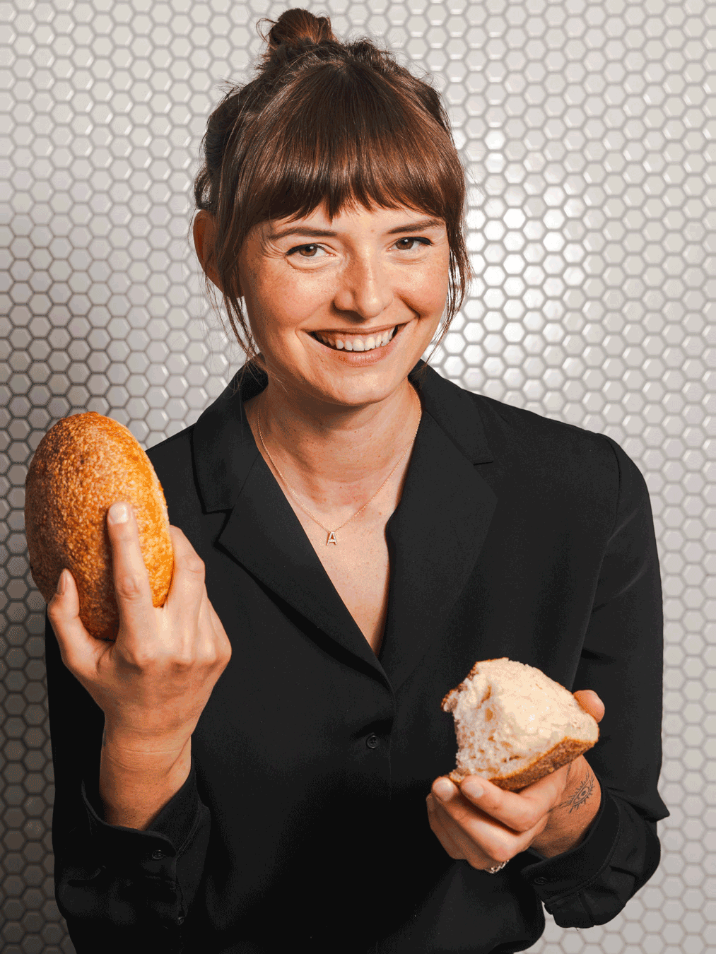 A woman with brown hair in a bun, wearing a black blazer, smiling, holding a bread roll in her left hand and a piece of bread in her right hand, standing against a textured background.